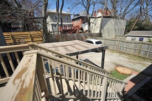Une terrasse en bois avec une rambarde en métal, une voiture blanche garée à l’arrière-plan et un quartier résidentiel avec des maisons et des arbres.