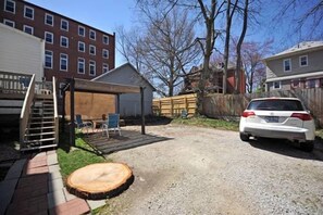 A residential backyard with a gravel driveway, a wooden deck, a covered patio, and a white car parked in the background. - Spacious 3BR Crescent Hill Apt | Yard & Parking (Louisville)