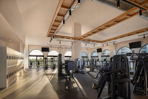 A spacious gym with multiple exercise machines, large windows, and a high ceiling with exposed beams.