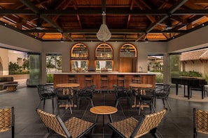 A bar area with a central counter, surrounded by wicker chairs and tables, with a view of the outdoors.