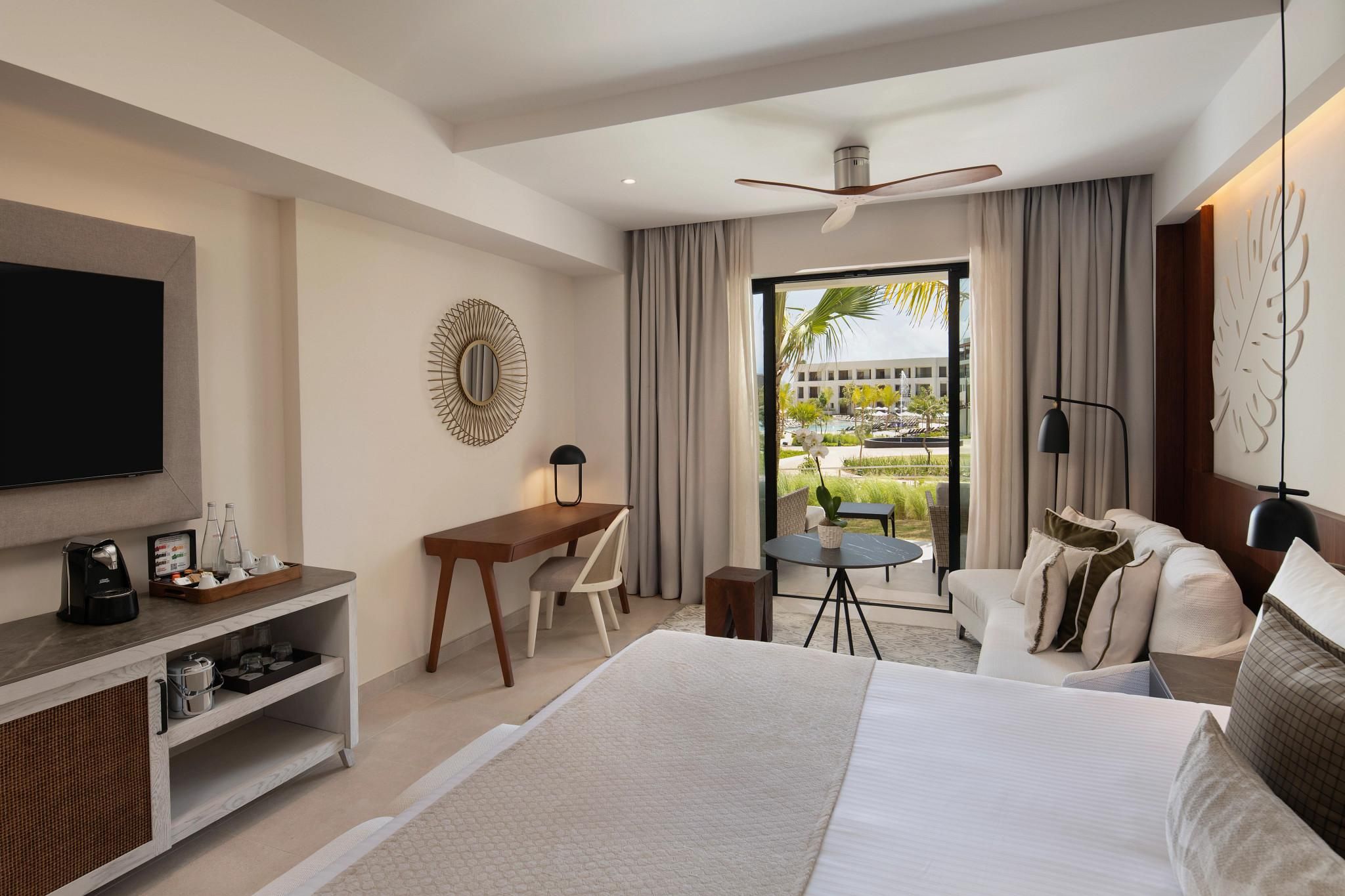 A hotel room with a bed, a desk, a TV, a chair, a small table, and a view of a pool and buildings.