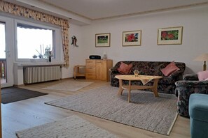 A living room with a sofa, a coffee table, a radiator, a window with curtains, and framed pictures on the wall. - Holiday apartment with 2 bedrooms (Sasbachwalden)