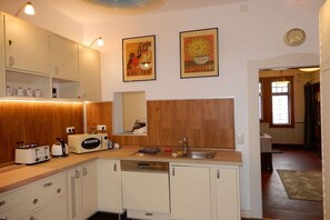 A compact kitchen with white cabinetry, a wooden countertop, and a built-in dishwasher. There are two wall-mounted posters, one with a bird and coffee theme and another with a sun and coffee theme. A clock is visible on the wall. - Holiday home with 4 bedrooms (Hundsdorf)
