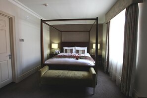 Una cama con dosel, mesitas de noche con lámparas, un banco al pie de la cama y vistas al exterior a través de cortinas.