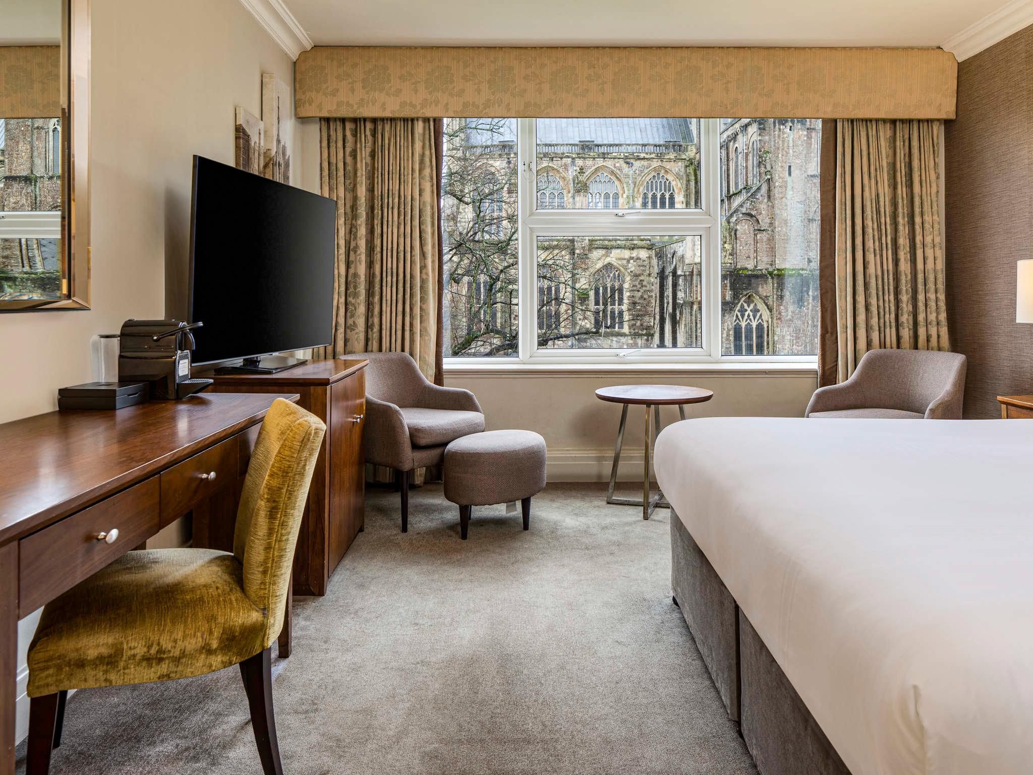 A hotel room with a bed, a desk, a chair, a television, and a view of historic buildings through the window.