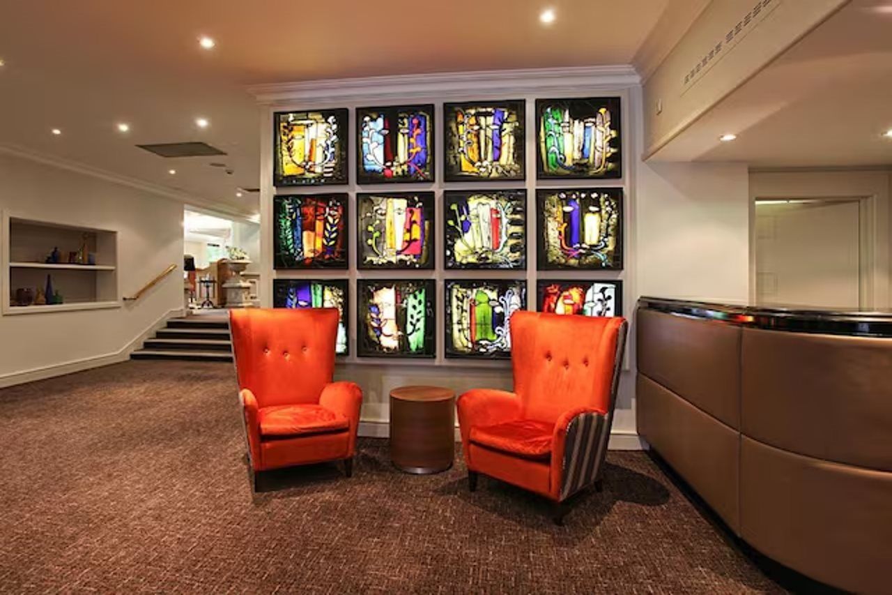 A lobby with a wall of stained glass windows, two orange armchairs, a small round table, and a curved reception desk.