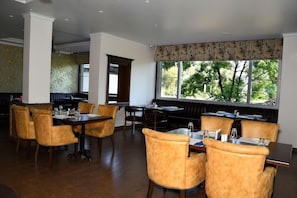 Un restaurante con vistas a árboles a través de amplios ventanales, mesas y sillas de madera, y una pared con diseño.