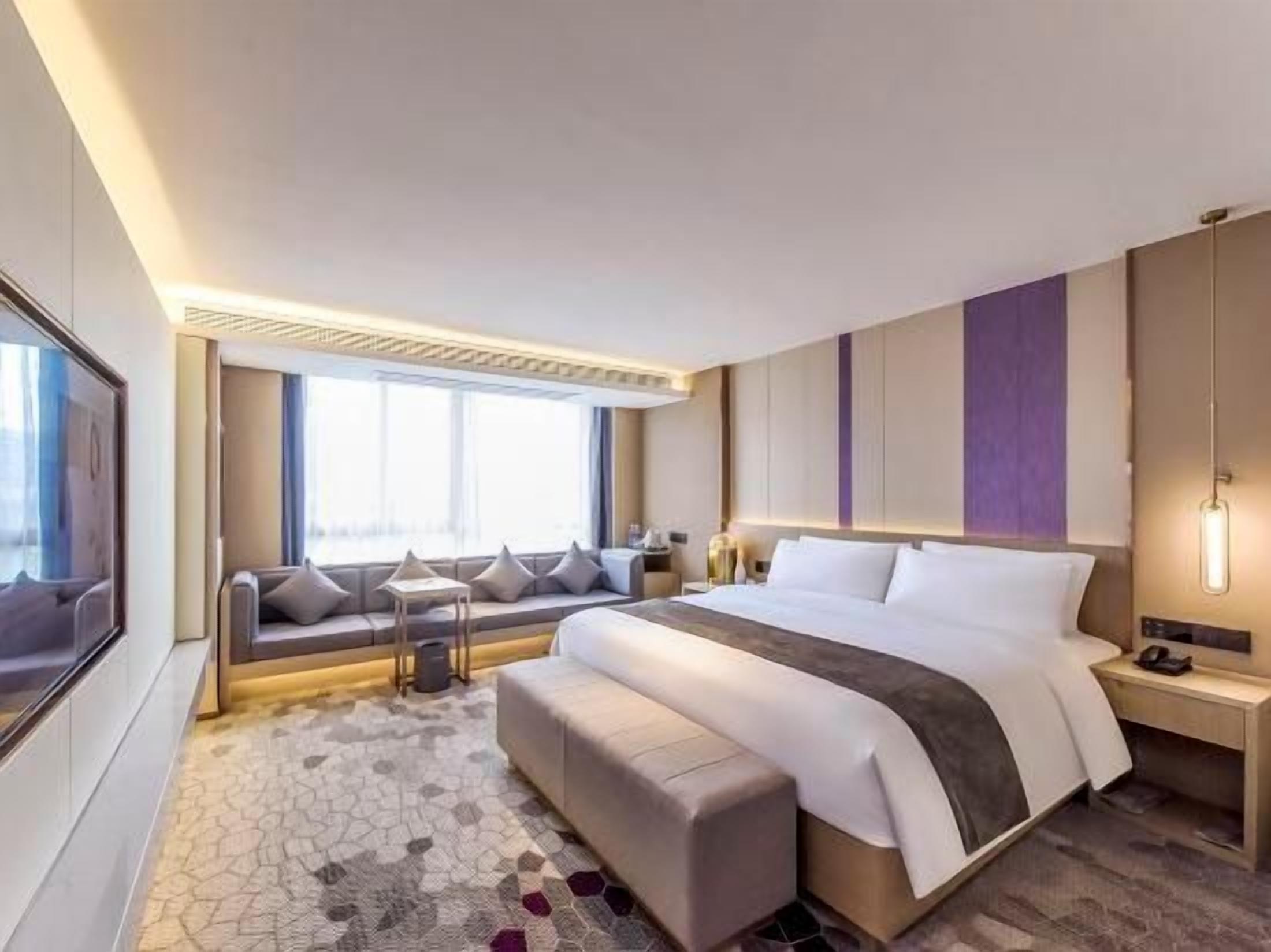 A modern hotel room with a large bed, a seating area with cushions, a desk, and a large window.