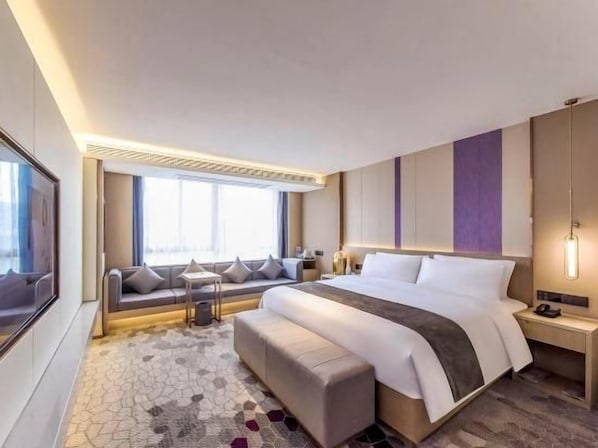 Quarto de hotel moderno com uma cama grande, um espaço de estar com almofadas, uma escrivaninha e uma janela ampla.