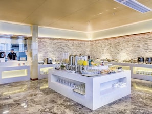 Disposição de buffet com piso de mármore, área central de serviço com diversos pratos e uma pessoa ao fundo.