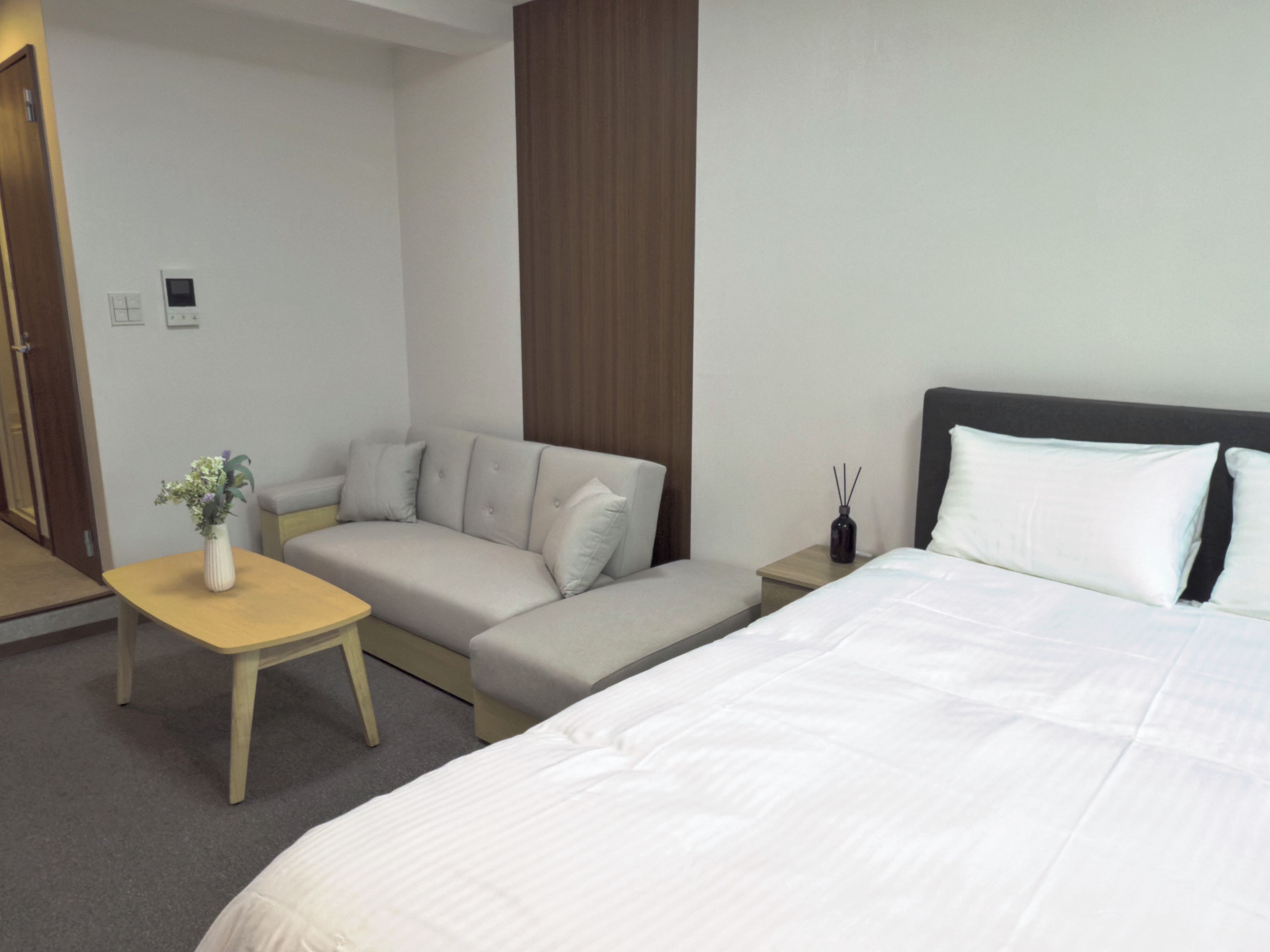 A hotel room with a bed, a sofa, a small table with a vase, and a nightstand with a bottle.