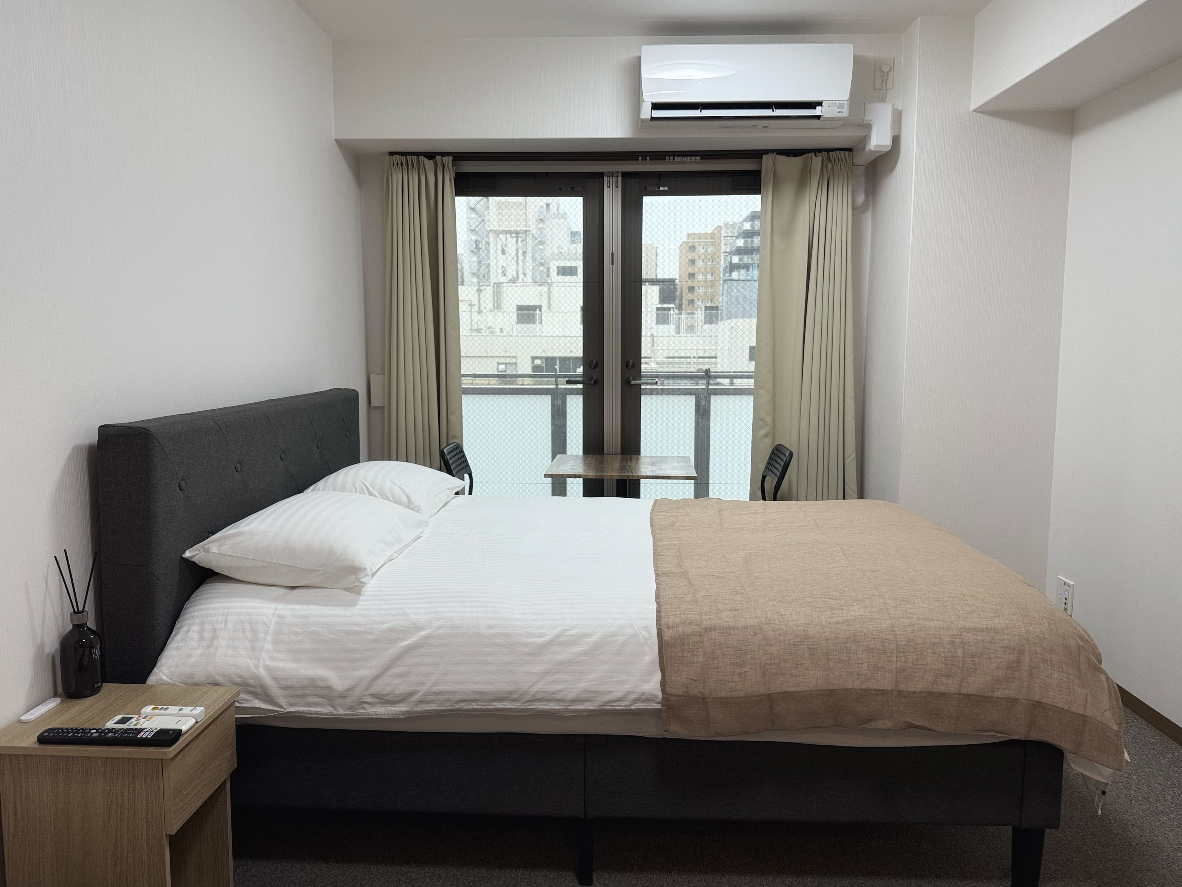 A hotel room with a bed, a bedside table with a remote control, a window with curtains, and an air conditioning unit.