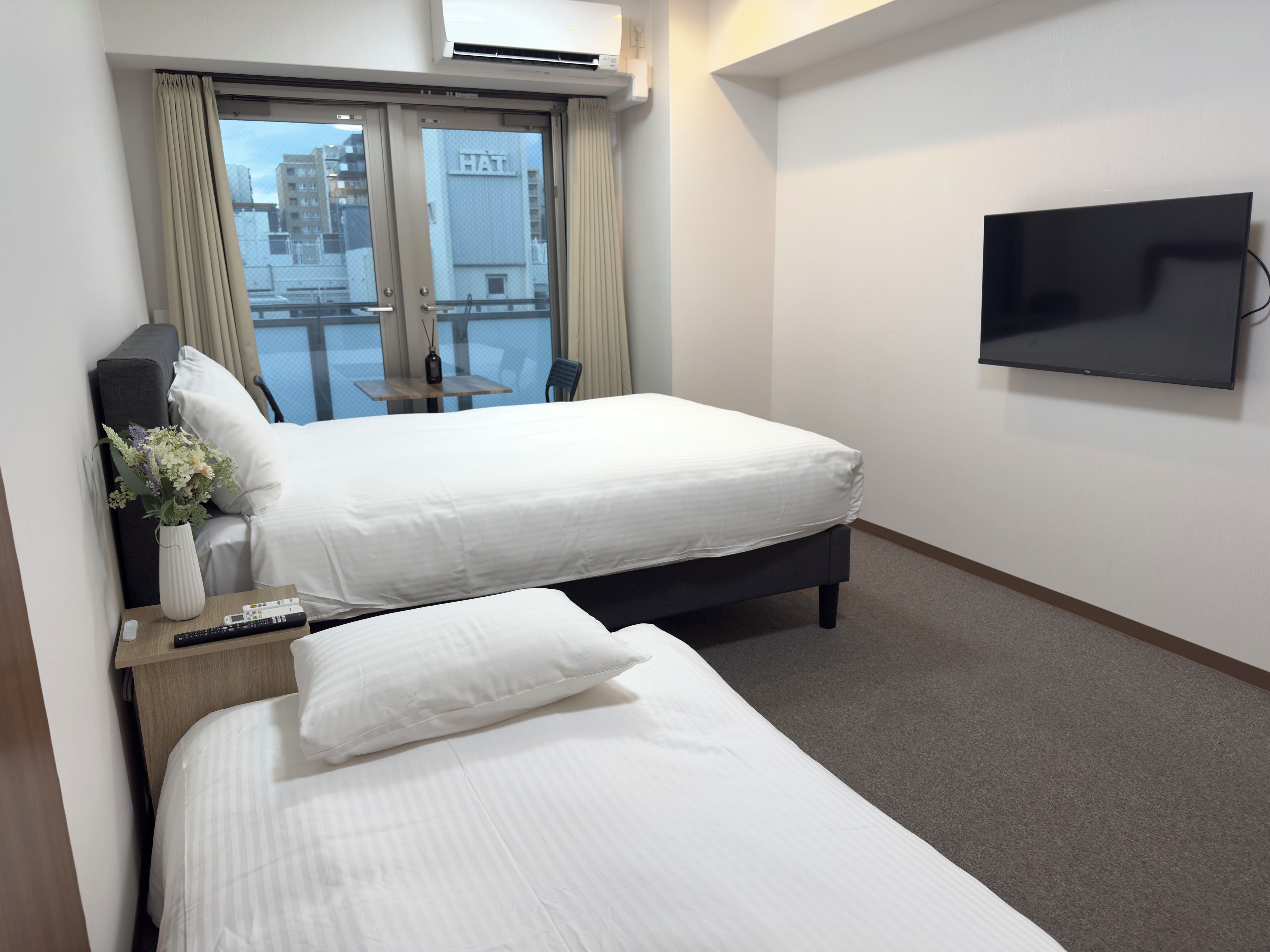 Habitación de hotel con dos camas, un televisor, una mesa con un florero y vistas a la ciudad desde la ventana.