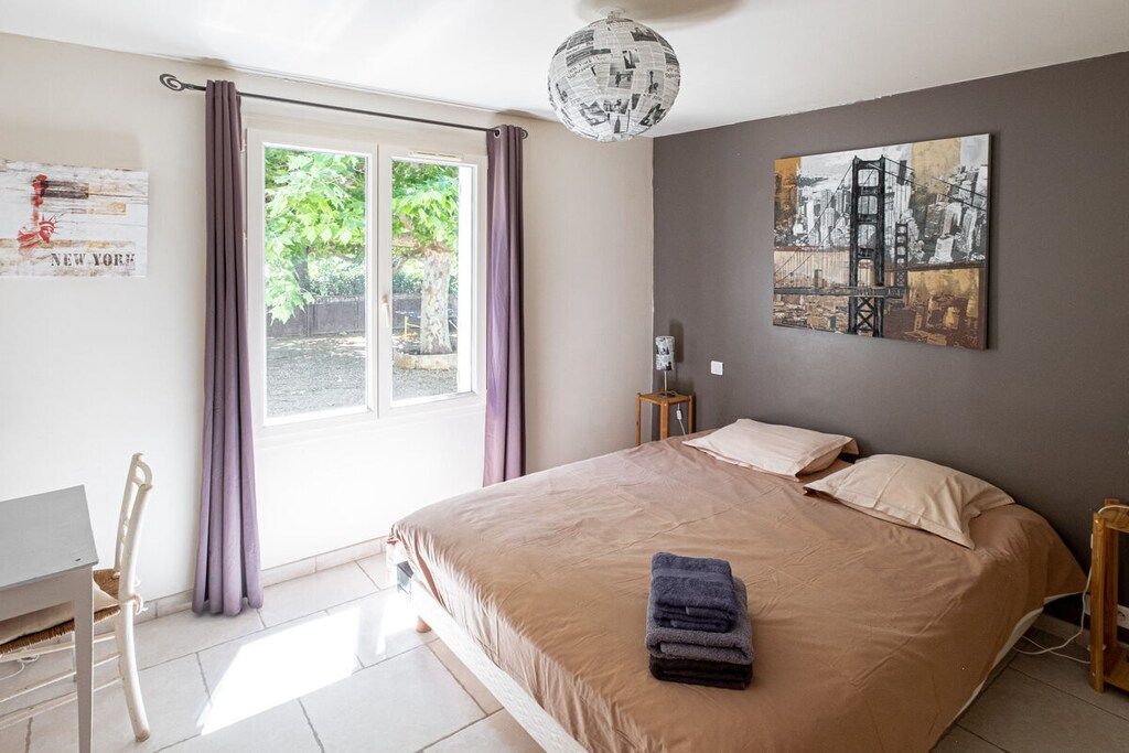 A bedroom with a bed, a window with curtains, a wall with a painting, and a chair.