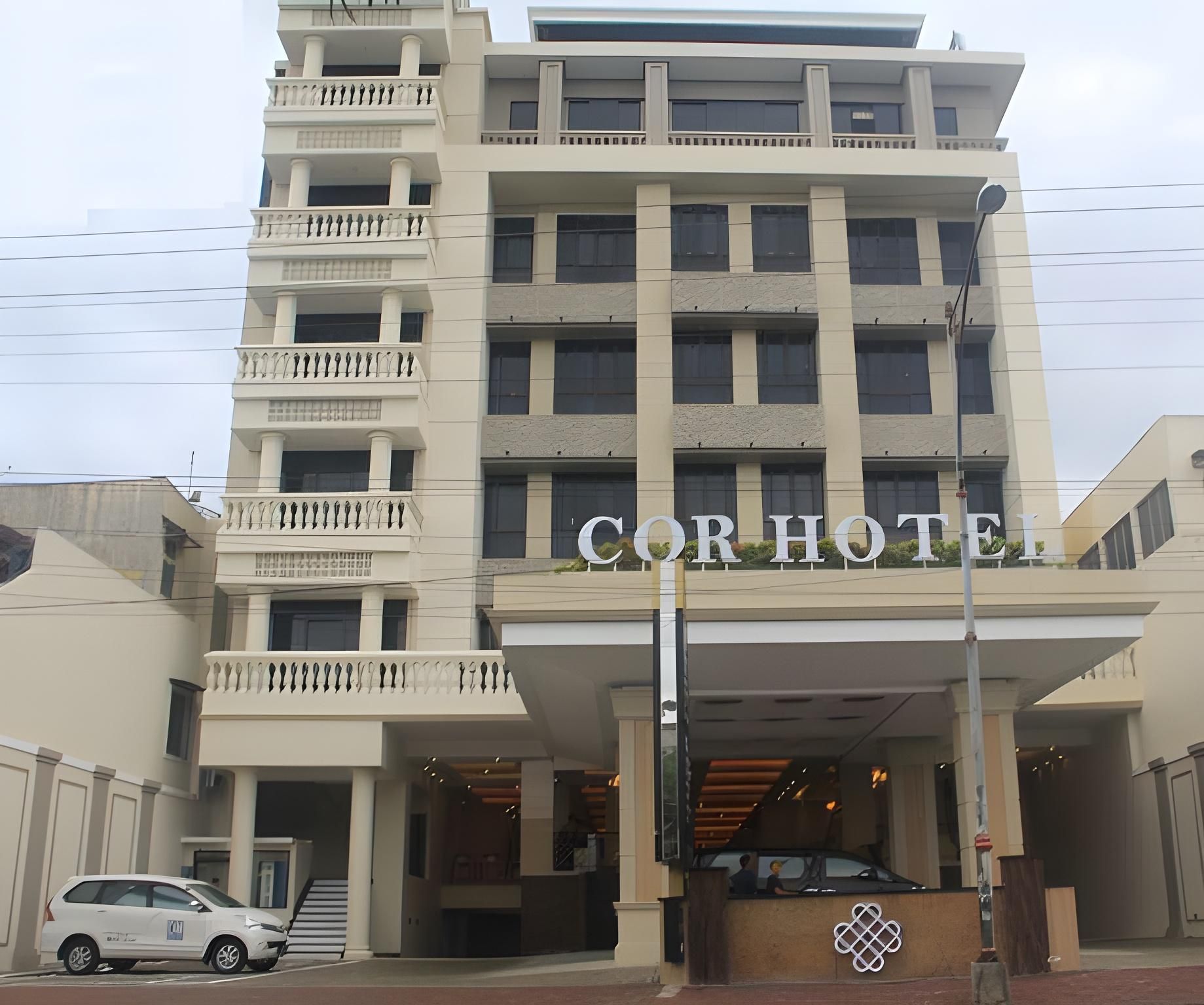 A multi-story hotel with a prominent 'CORHOTEL' sign, a covered entrance, and a car parked in front.