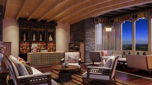 Amplia sala de estar con techo alto, pared de piedra y vistas por ventanas grandes. Hay varias zonas de estar con sofás y sillones, además de un aparador decorativo con estatuas.