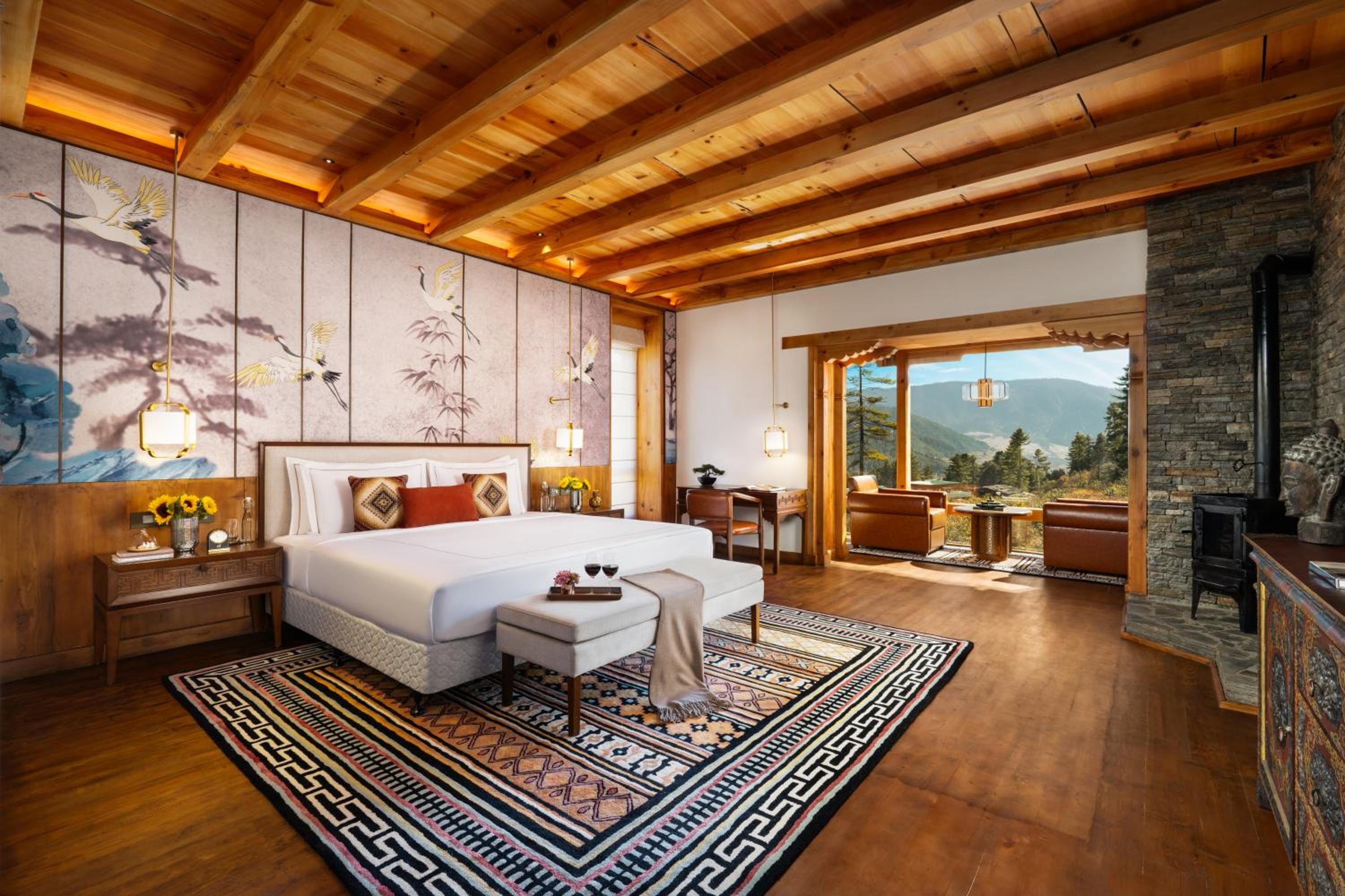 A spacious bedroom with a large bed, a wooden ceiling, a stone fireplace, and a view of the mountains.