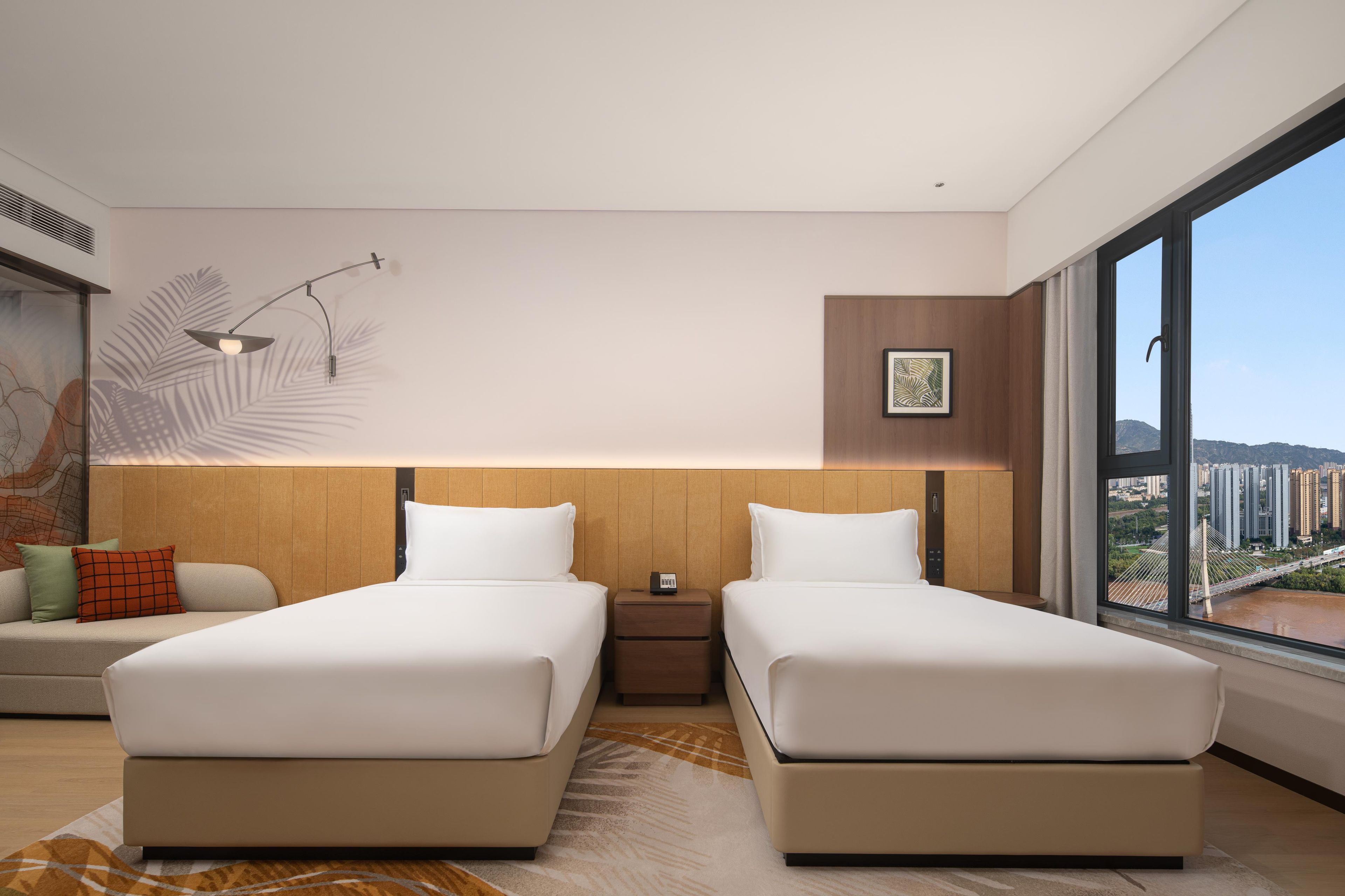 A modern hotel room with two beds, a nightstand, a large window with a city view, and a wall-mounted lamp.