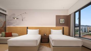 A modern hotel room with two beds, a nightstand, a large window with a city view, and a wall-mounted lamp.