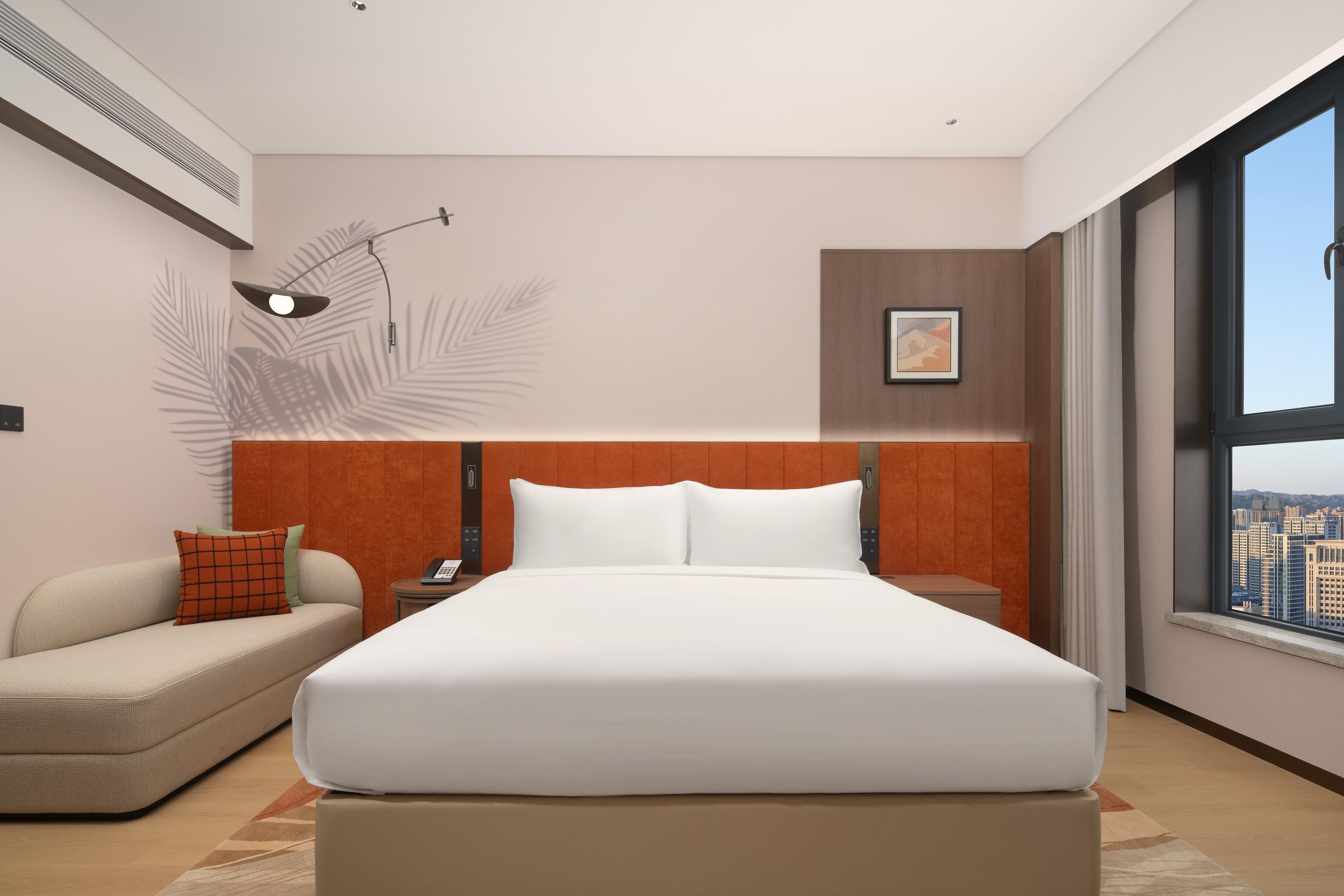 A modern hotel room with a large bed, a sofa, a nightstand, a wall-mounted lamp, and a city view.
