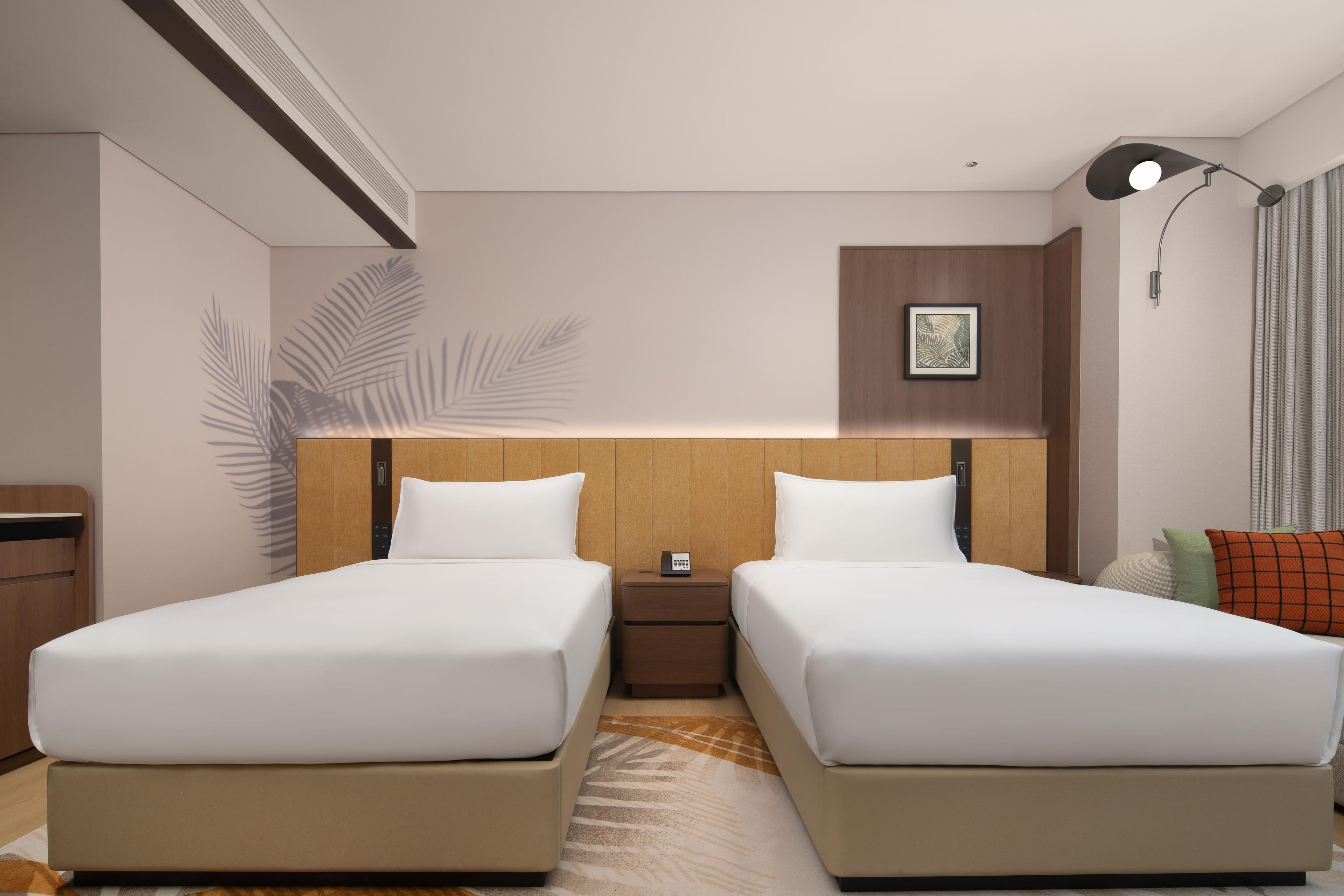 A hotel room with two beds, a nightstand, a wall-mounted picture, a lamp, and a window with curtains.
