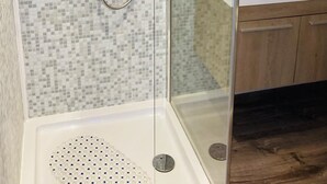Une douche fermée par des parois de verre, un mur recouvert de carreaux, un lavabo avec un miroir et des meubles en bois.