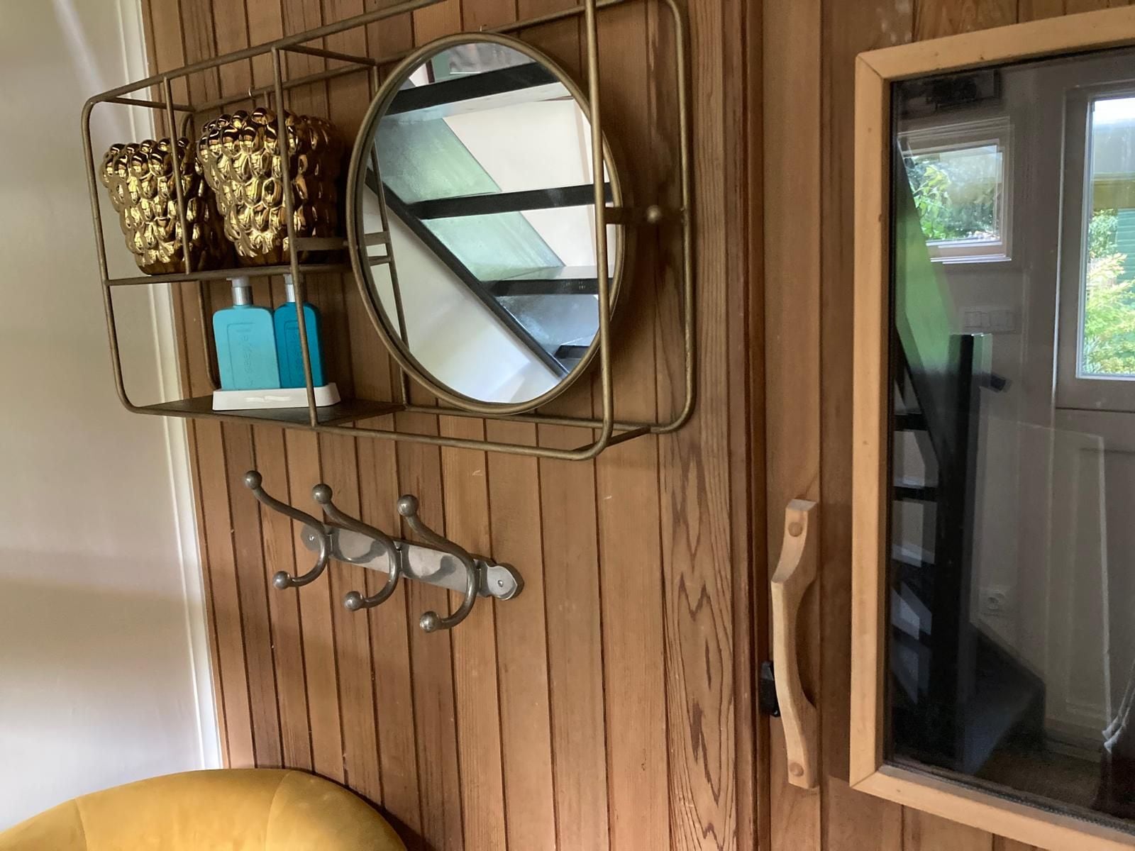 Un mur en bois sur lequel est fixée une étagère en métal où sont disposés des objets décoratifs, un miroir rond et une porte dotée d’une poignée.