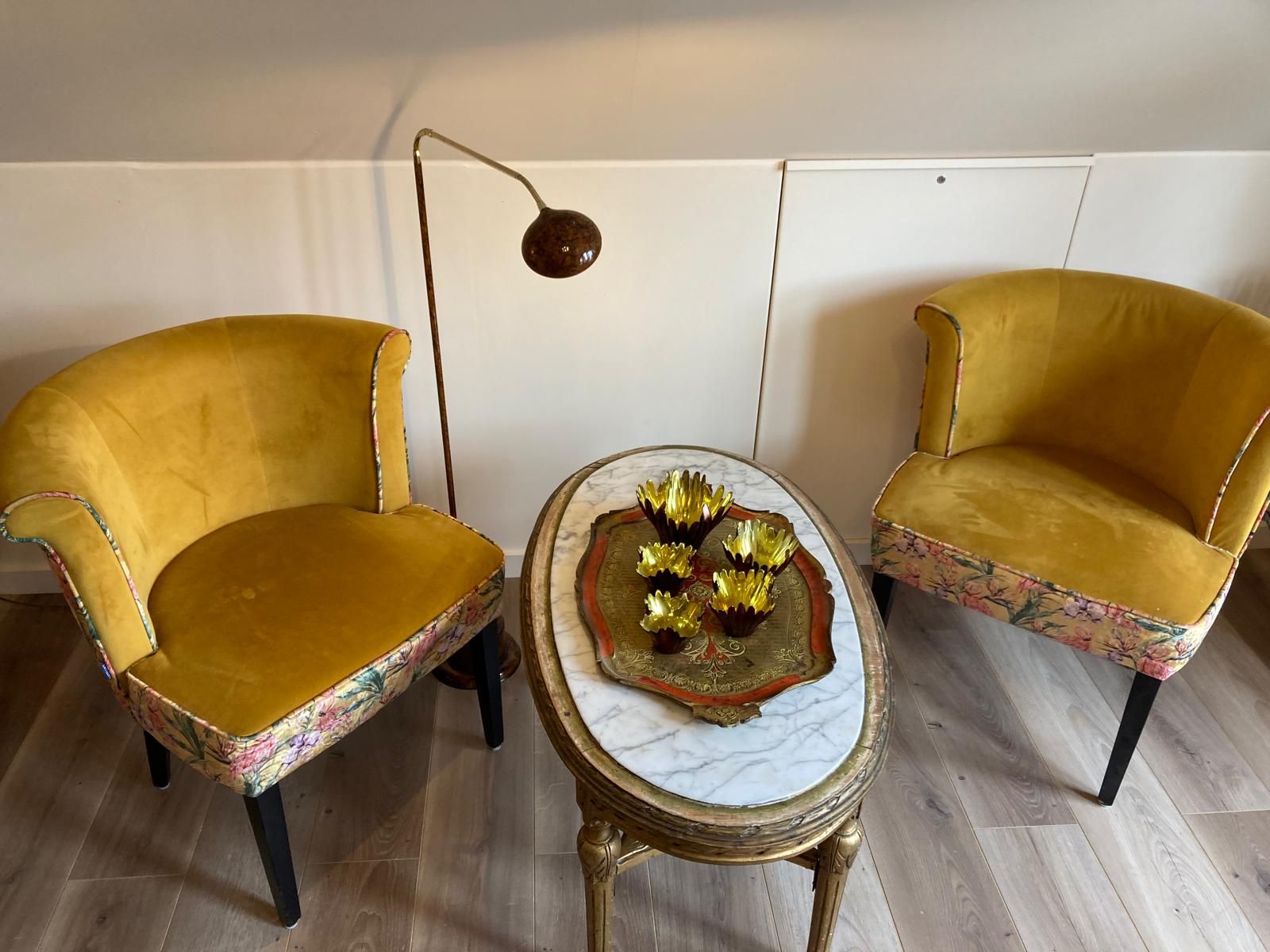 Duas poltronas com estofamento floral, uma mesa redonda pequena com tampo de mármore e um abajur fixado na parede.