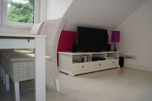 Quarto com aparador branco, televisão de tela plana, cadeira branca e janela com vista para área verde.