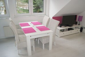 Ein weißer Esstisch mit rosa Platzsets, Korbstühle, eine weiße Kommode mit einem Flachbildfernseher und ein Fenster mit Blick auf Bäume.