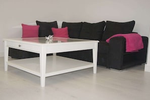Ein weißer Tisch mit Glasplatte, vor einer schwarzen Couch mit rosafarbenen Kissen platziert.