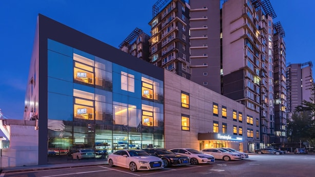 Un hotel moderno con fachada de vidrio, varios pisos y un estacionamiento con varios automóviles.