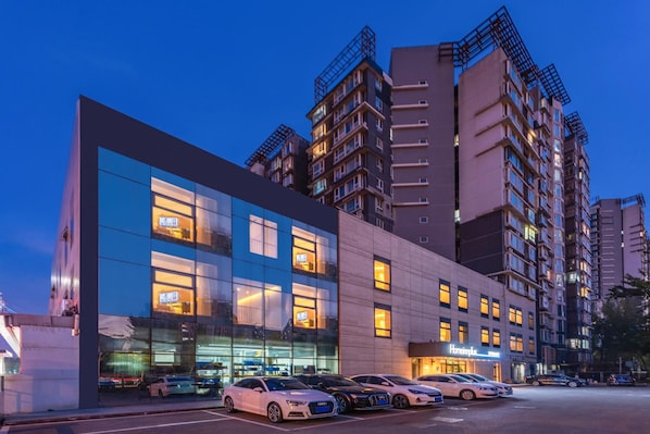 A modern hotel with a glass facade, multiple floors, and a parking lot with several cars. - Homeinn Plus (Beijing)