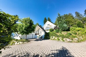 A residential property with a paved driveway, a white house with a gabled roof, and a surrounding landscape with trees and shrubs. - Holiday home with 2 bedrooms (Sasbachwalden)