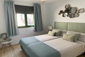Ein Schlafzimmer mit einem Bett, zwei Kissen, einem Sessel, einem Fenster mit Vorhängen und einem Wandspiegel.