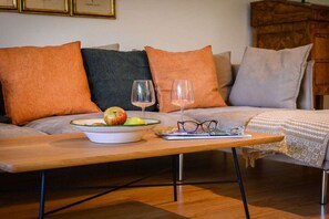 Ein gemütliches Wohnzimmer mit einer Couch, Kissen, einem Holztisch, einer Obstschale, einem Weinglas und einem Brillenpaar.