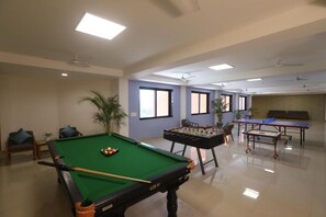 A pool table with a green felt surface, a foosball table, and a ping pong table in a room with a high ceiling and large windows.