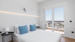 A modern bedroom with a large bed, white bedding, two blue patterned pillows, a bedside table with a lamp, and a view of the outside through a sliding glass door. - Celine: Apartments in the Center (Thessaloniki)