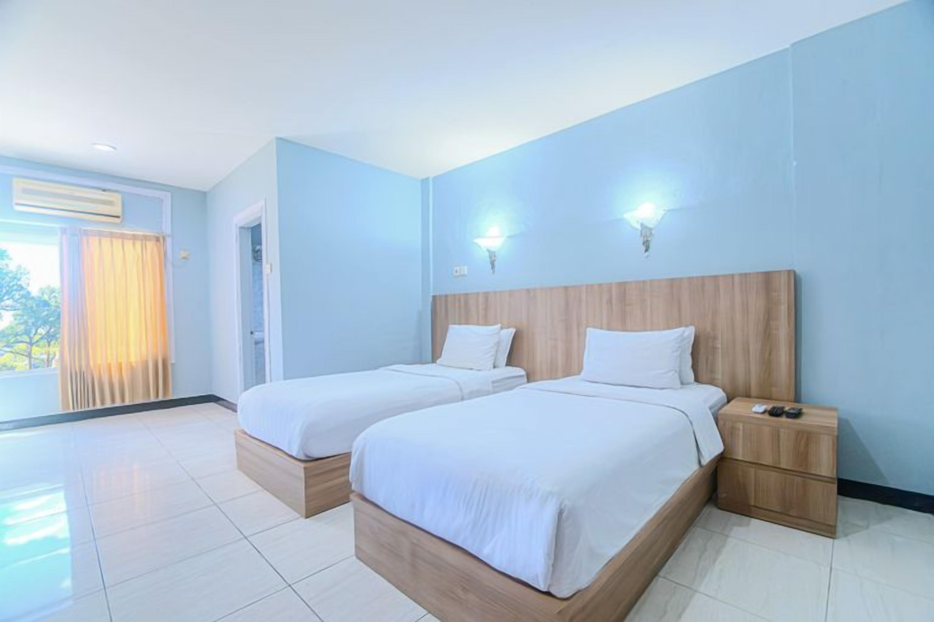A hotel room with two beds, a wooden headboard, a nightstand, and a view of the outdoors.