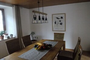 Een eethoek met een houten tafel, stoelen, een fruitschaal, een hanglamp en een ingelijste botanische afbeelding aan de muur.