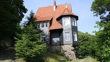Ein zweigeschossiges Haus mit rotem Ziegeldach, Steinunterbau und umgeben von Grün.