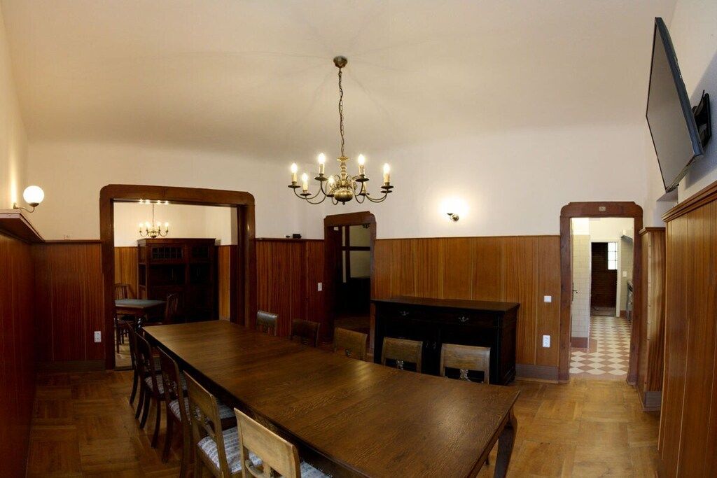 A dining area with a long wooden table, chairs, a chandelier, and wooden paneling on the walls.