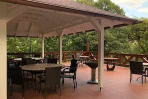 Eine Außenpatio mit Tischen und Stühlen, einem Grill und einer Holzterrasse mit Blick auf einen bewaldeten Bereich.