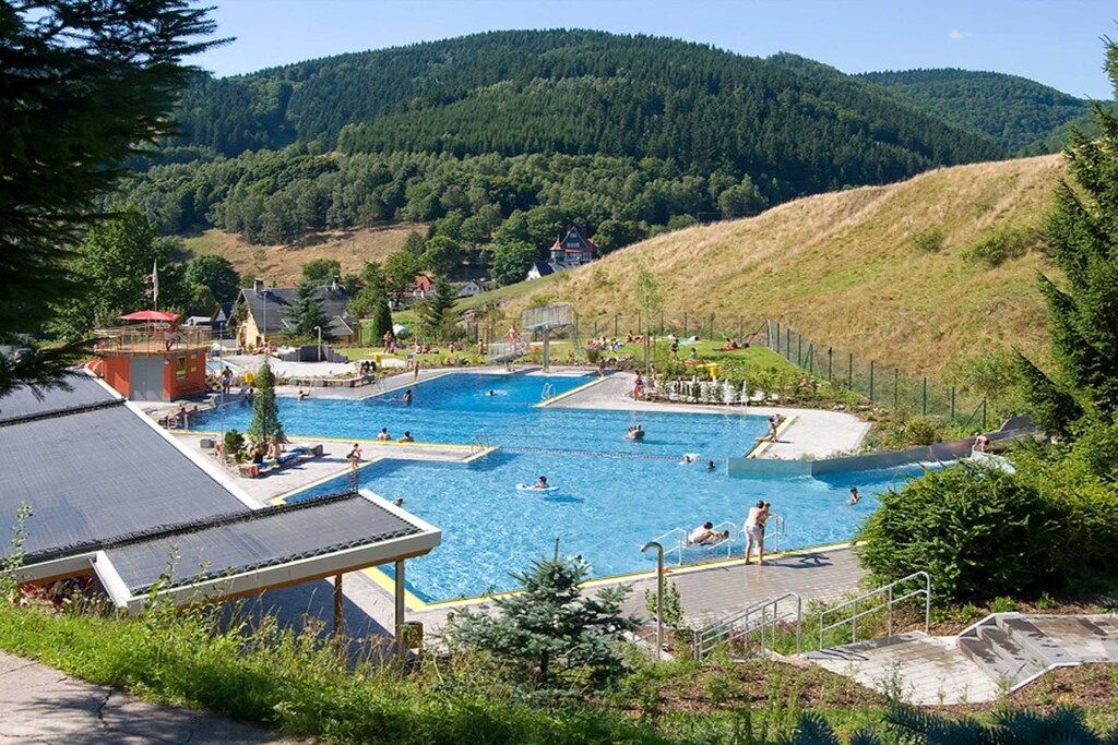 Ein Schwimmbadkomplex mit mehreren Pools und einer umlaufenden Liegefläche, vor einer Kulisse aus Hügeln und Bäumen.