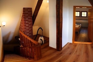 Eine Holztreppe mit Kamin aus Ziegelsteinen, ein Staubsauger und ein Bett in einem Raum mit Holzboden und Heizkörper.