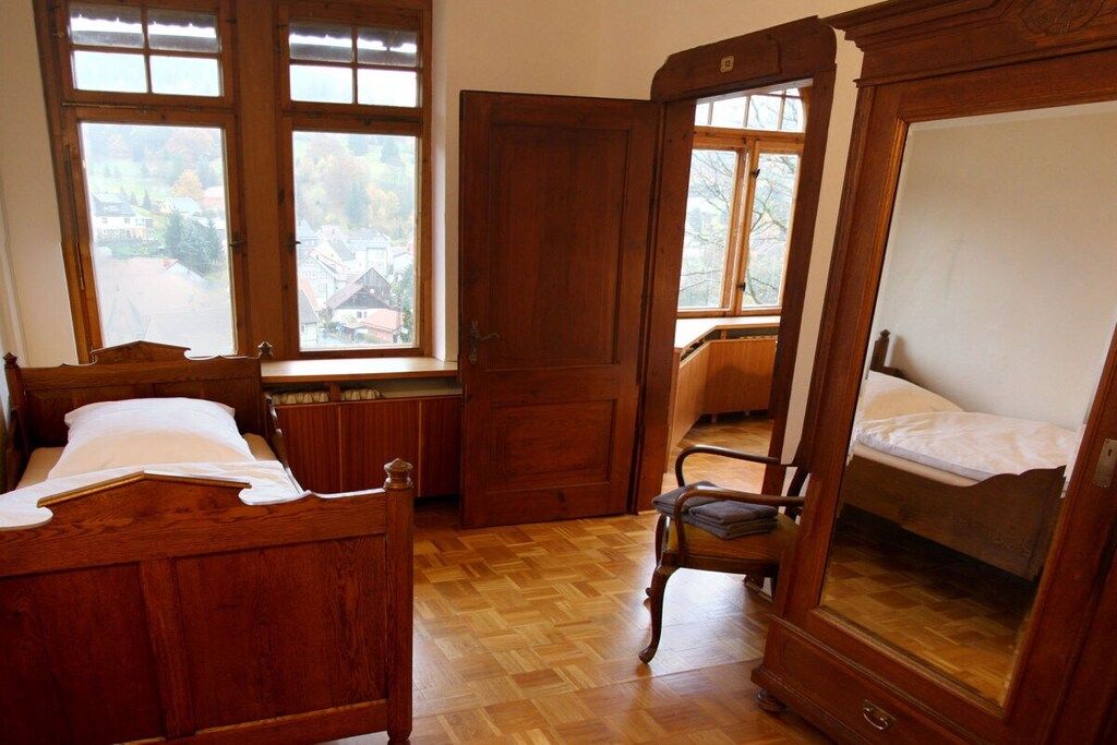 A room with a wooden bed, a chair, and a window with a view of houses.