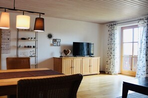 A living room with a wooden dining table, wicker chairs, a television on a wooden cabinet, and a window with curtains. - Apartment AusZeit in Finning (Finning)