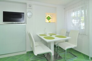 A small, clean room with a TV, a table with chairs, a window with curtains, and a green tiled floor. - Spacious modern apartment with big patio and sea view (Slatine)