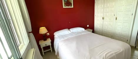 Een slaapkamer met een bed, een nachtkastje, een lamp, een schilderij en een kledingkast.
