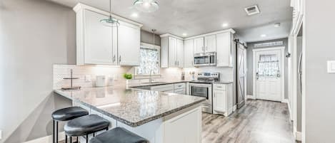 A modern kitchen with a large island, bar stools, stainless steel appliances, and a herringbone-patterned backsplash.